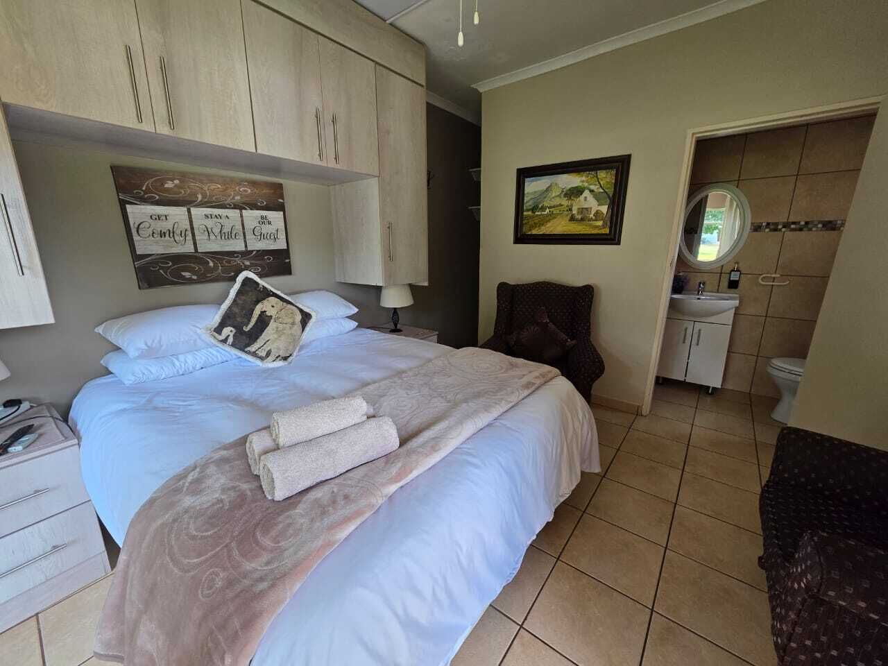 A bedroom with a bed, a nightstand, a dresser, a painting, and a bathroom visible through an open door.