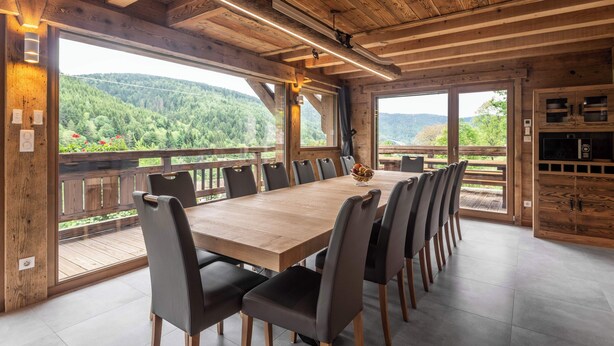 A wooden dining area with a large table and chairs, large windows with a view of a forested area.
