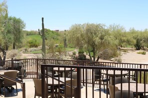 An outdoor patio with wicker furniture, a view of a landscaped area with trees and a cactus, and a distant building.
