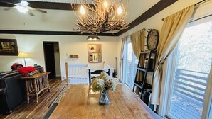 Uma sala de jantar com mesa de madeira, cadeiras pretas, um candelabro e vista para o exterior através de grandes janelas.