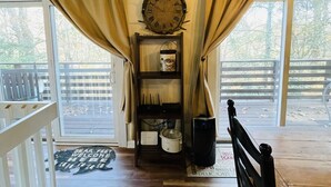 Quarto com piso de madeira, um relógio na parede, prateleira com utensílios de cozinha e um banco.