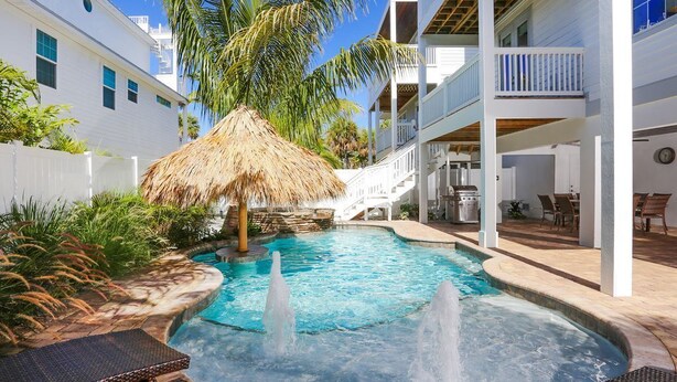 A swimming pool with two water jets, surrounded by lounge chairs, a thatched umbrella, and a white building with balconies.