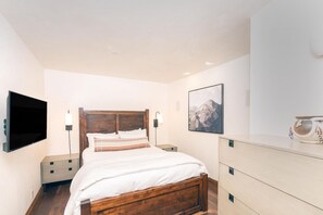 A bedroom with a wooden bed, bedside tables, a flat-screen TV, a wall-mounted lamp, and a framed picture.