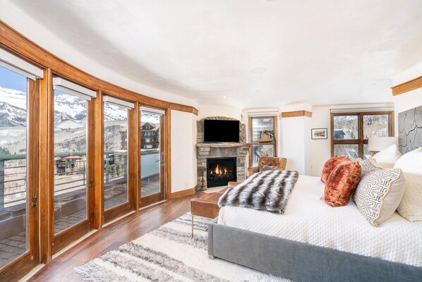 Un dormitorio espacioso con una cama grande, una chimenea y vistas a un paisaje montañoso nevado a través de ventanales de piso a techo.