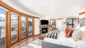 A bedroom with a fireplace, a bed, a sofa, and a view of a snowy landscape through large windows. - Penthouse At The Peaks 623 (Mountain Village)