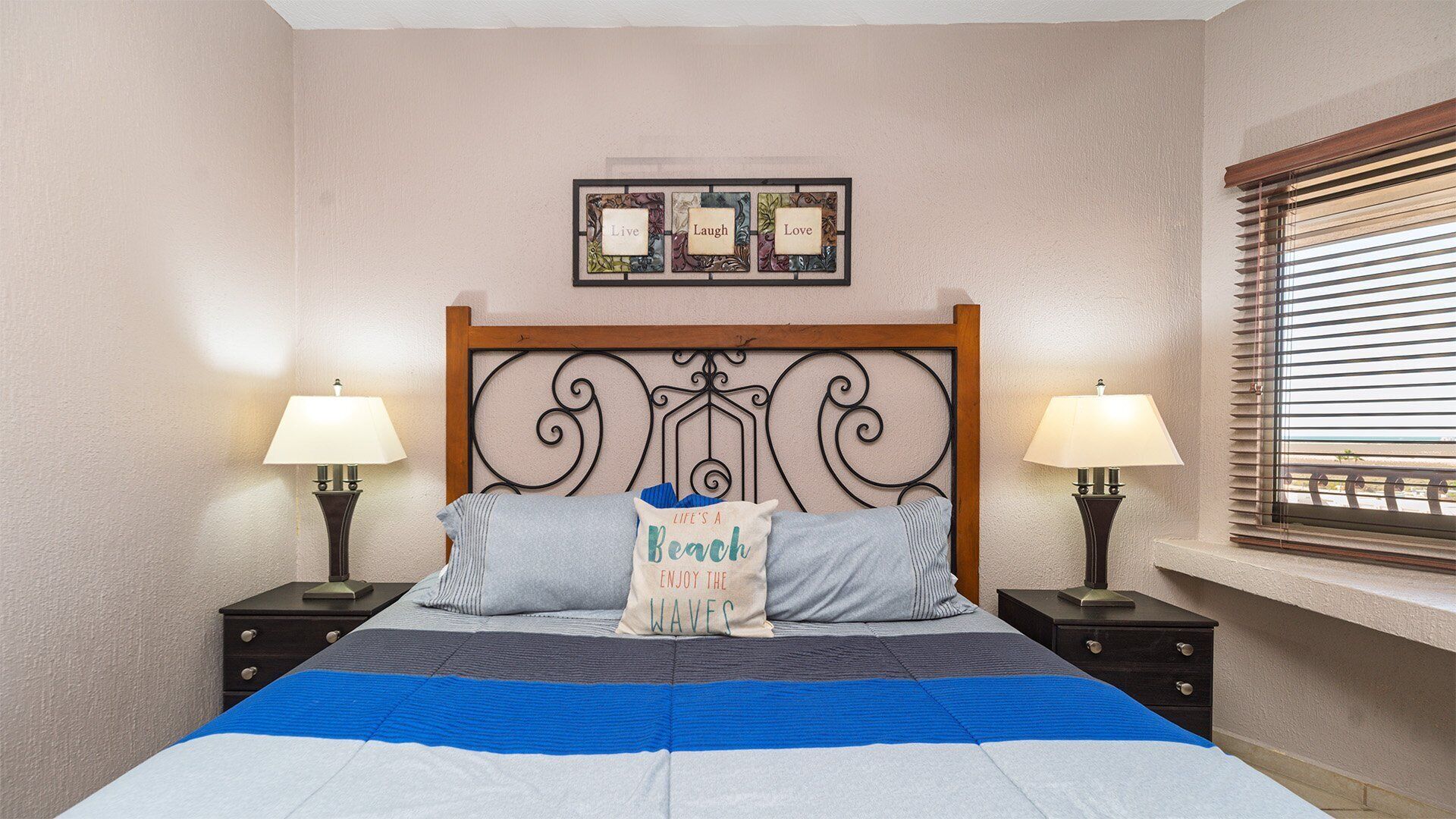 A bedroom with a bed, two bedside tables with lamps, a wall-mounted picture frame, and a window with blinds.