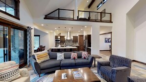 A spacious living area with a dark gray sofa, a patterned armchair, and a coffee table. A kitchen with modern appliances and a dining area with a large table and chairs is visible in the background. - Family Fun Lodge with Access to Exclusive Golf (Bend)