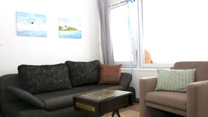 A living room with a dark grey sofa, a smaller armchair, and a small table with a decorative item on it. There are two paintings on the wall and a window with curtains. - Vacation Apartment K1213 with a View of the Baltic Sea (Schönberg OT Holm)