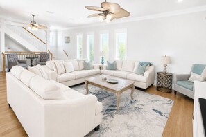 Ein geräumiges Wohnzimmer mit einem großen Ecksofa, einem Couchtisch und einem Kamin. Eine Treppe mit Geländer führt zu einer oberen Ebene.