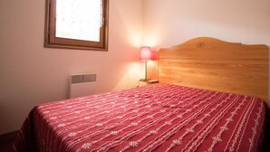 Un dormitorio con cabecera de madera, colcha estampada roja, lámpara de noche y ventana con persianas.