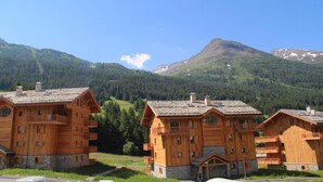 Una hilera de cabañas de madera con cimientos de piedra, frente a un bosque verde y una montaña.