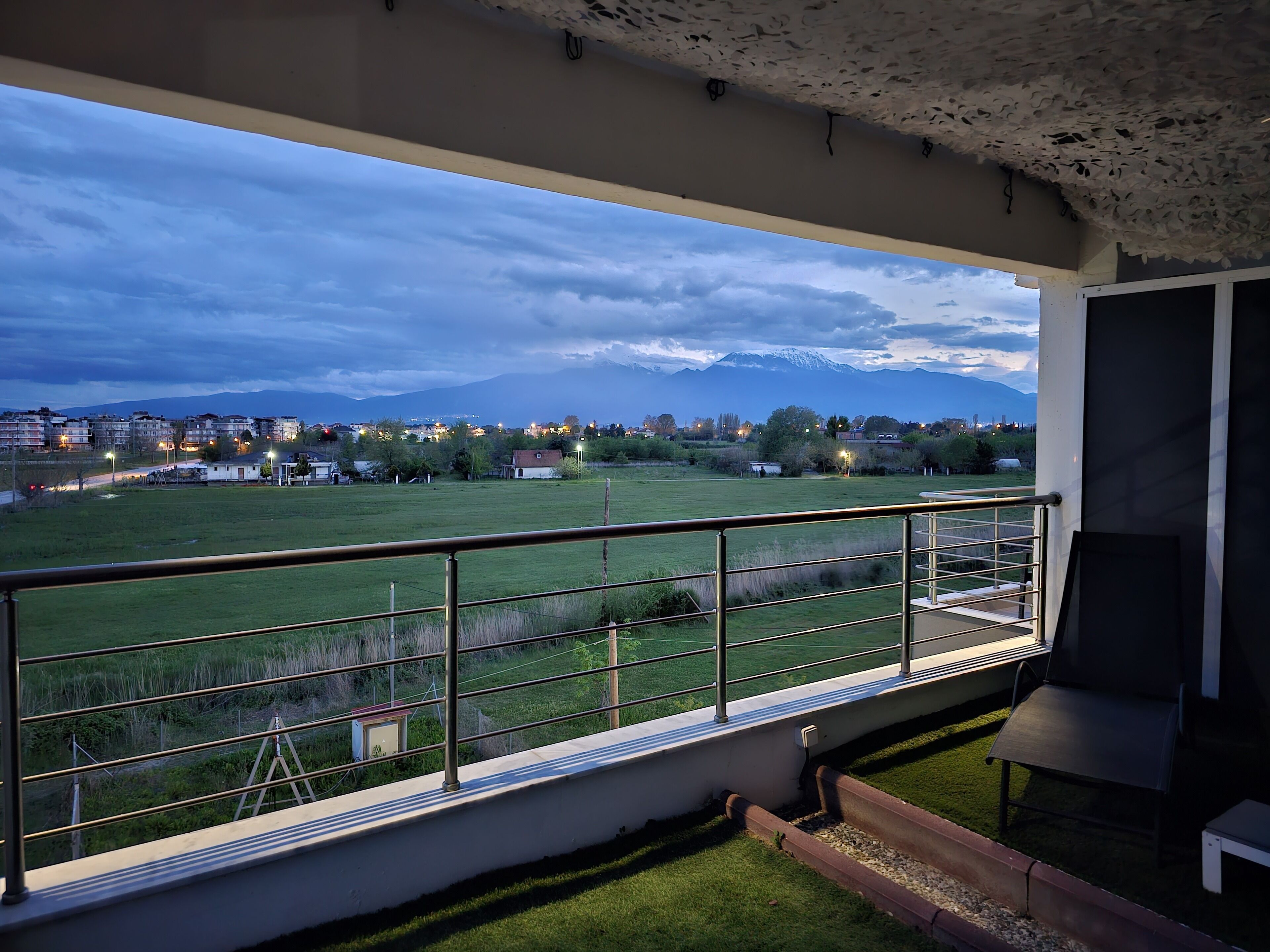 Uma varanda com vista para um campo verde e edifícios distantes, grades de metal e um espaço de estar pequeno com mesa e cadeiras.