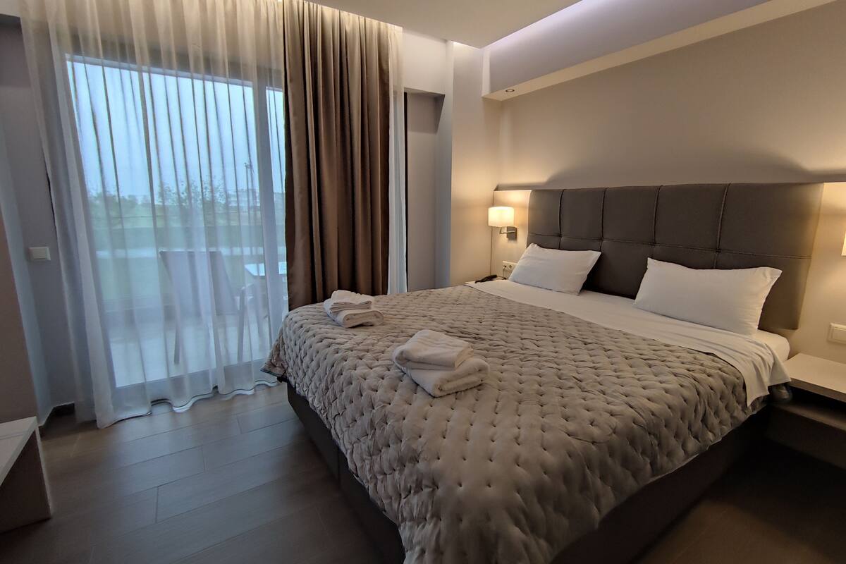 Habitación de hotel moderna con una cama grande, una mesita de noche, una lámpara de pared y vistas a través de cortinas translúcidas.
