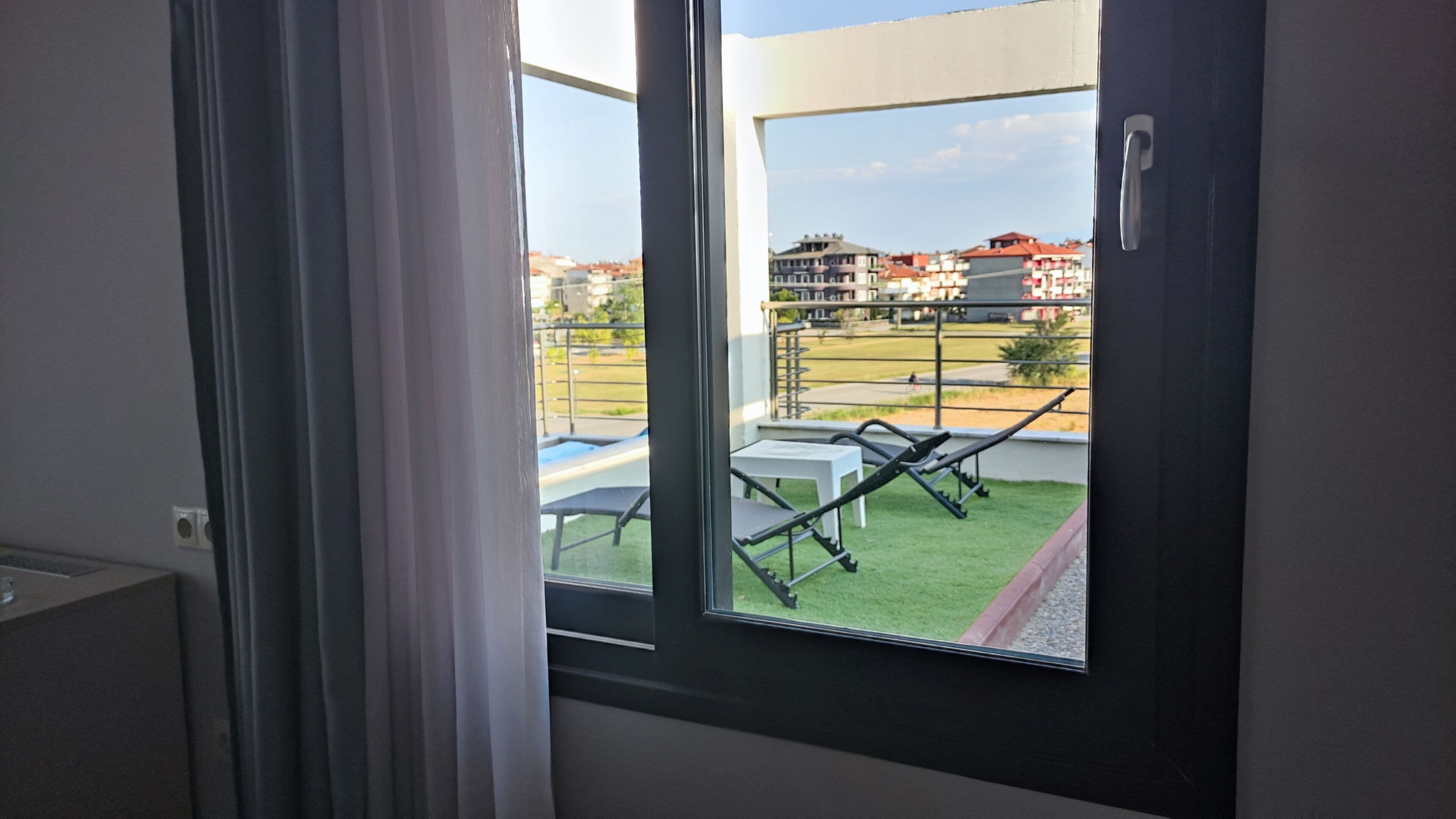 Une fenêtre donnant sur un balcon aménagé avec du mobilier d’extérieur et une pelouse.