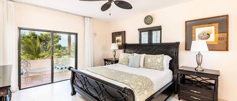 Un dormitorio con cama, ventilador de techo, mesita de noche con lámpara, cómoda y balcón con una silla.