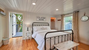 Un dormitorio con cama, una mesita de noche, un pequeño banco, una ventana con cortinas y un cartel que dice "COUNTRY HOME".