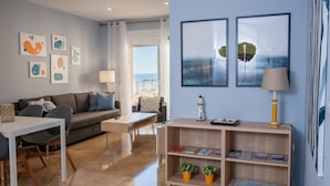 Una sala moderna con un sofá, una mesa de comedor y una estantería. Hay pinturas abstractas en la pared y una vista al mar a través de una ventana.