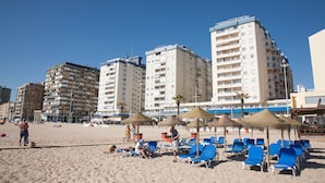 Una playa con sillas azules de descanso y sombrillas de paja, rodeada de altos edificios residenciales.