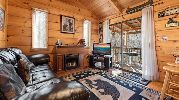 A cozy cabin interior with a leather couch, a fireplace, a television, and a deck with a glass door. - Forever Mine: A Romantic Escape for Two (Gatlinburg)