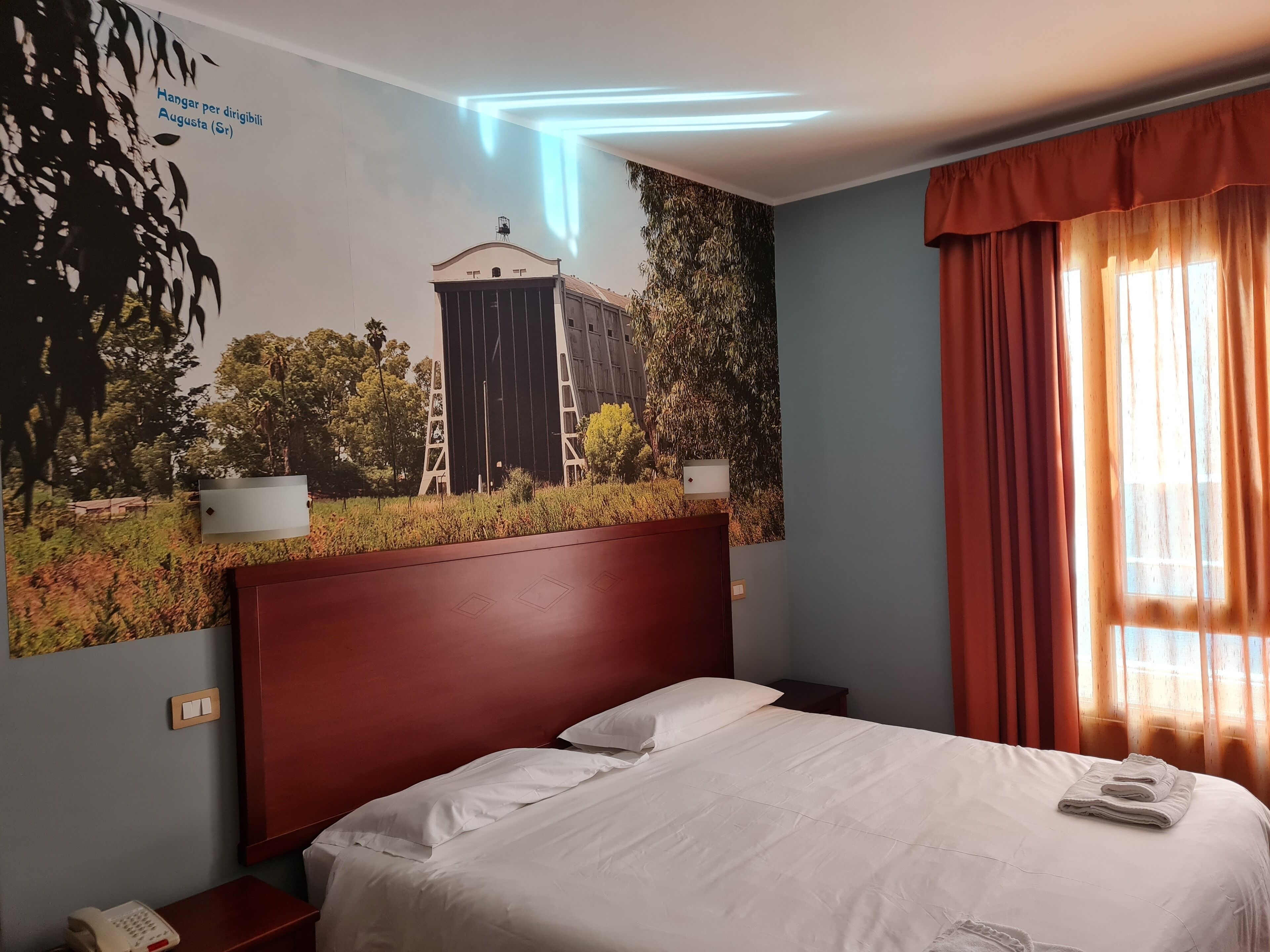 A hotel room with a bed, a nightstand, a wall-mounted telephone, and a window with curtains.