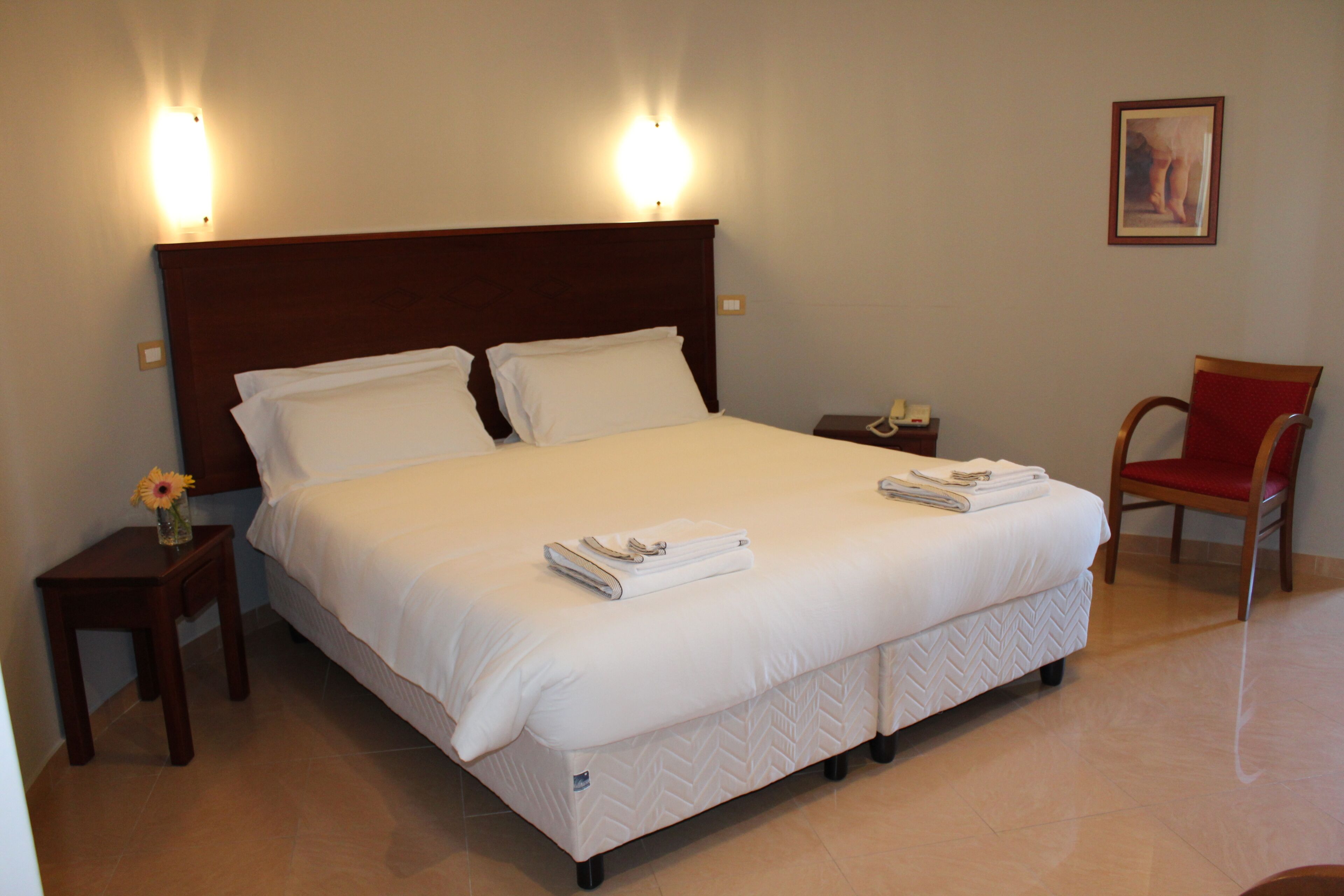 A neatly made bed with white linens, two bedside tables with lamps, a chair, and a framed picture on the wall.