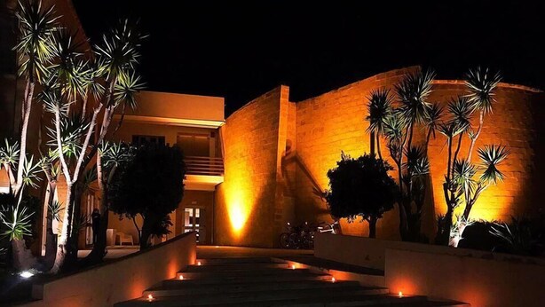 A building with a modern architectural design, featuring a prominent entrance with steps and a well-lit pathway. - Città Della Notte (Melilli)