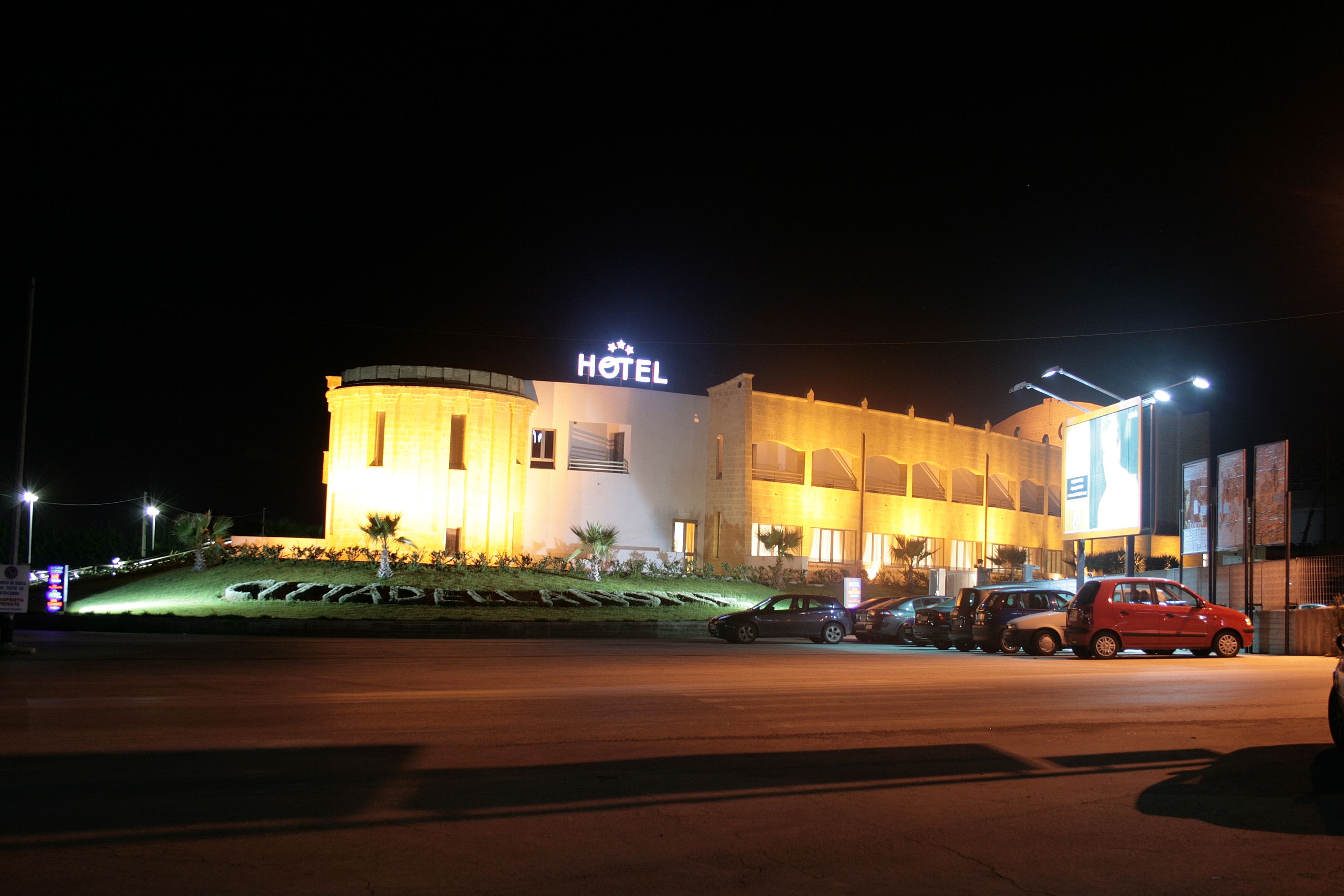 A hotel with a prominent "HOTEL" sign, a parking lot with several cars, and a well-lit facade.