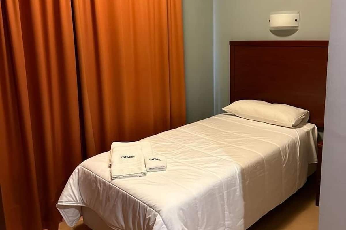 A hotel room with a bed, orange curtains, a headboard, and a wall-mounted light fixture.