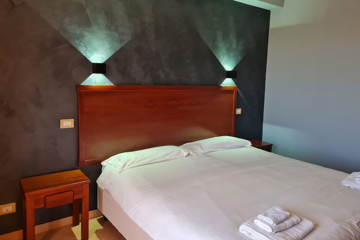 A neatly made bed with white linens and two pillows, a wooden nightstand with a lamp, and a wall with a textured finish and two wall-mounted lights.