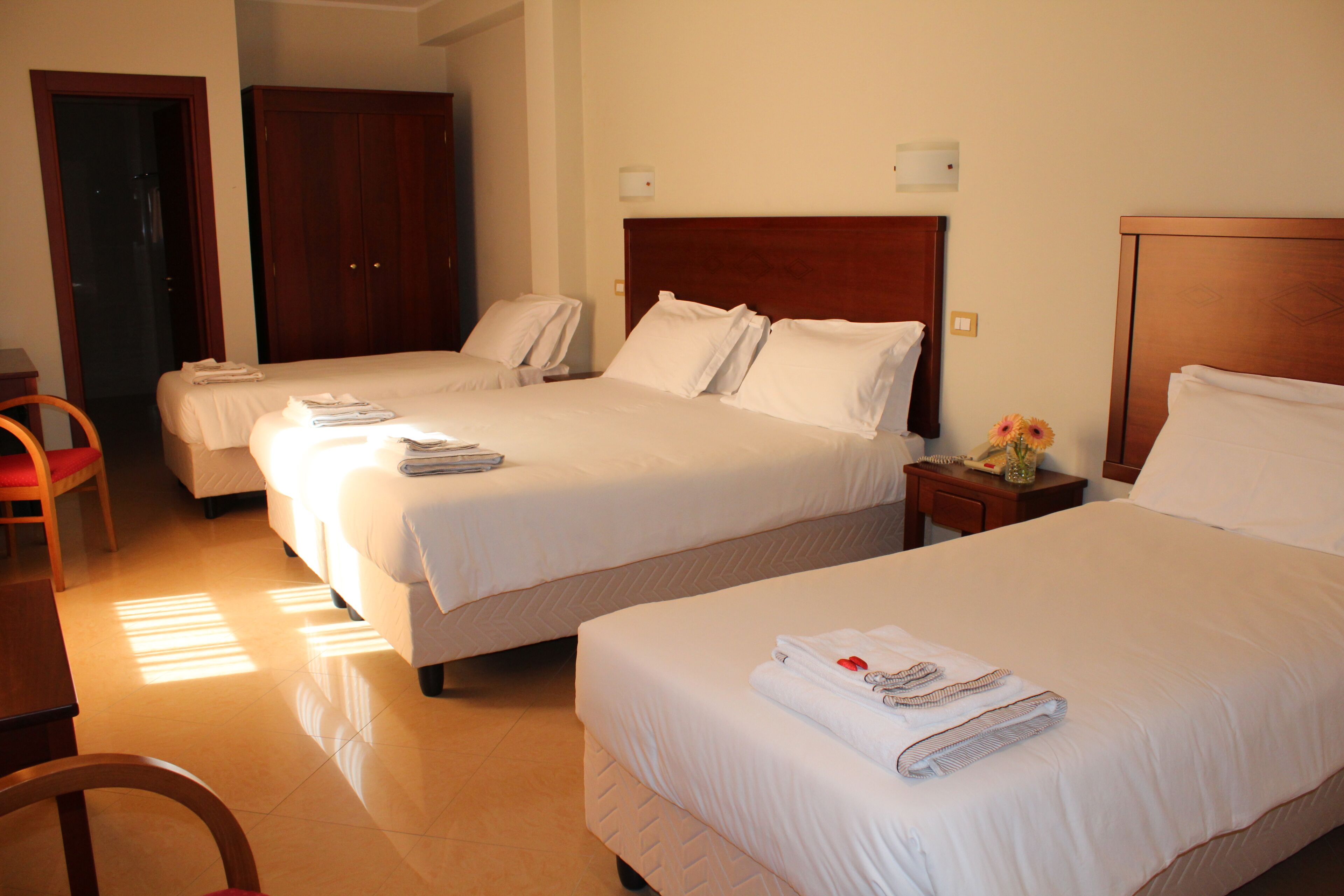 A hotel room with two beds, a wooden wardrobe, a small table with a vase, and a chair.