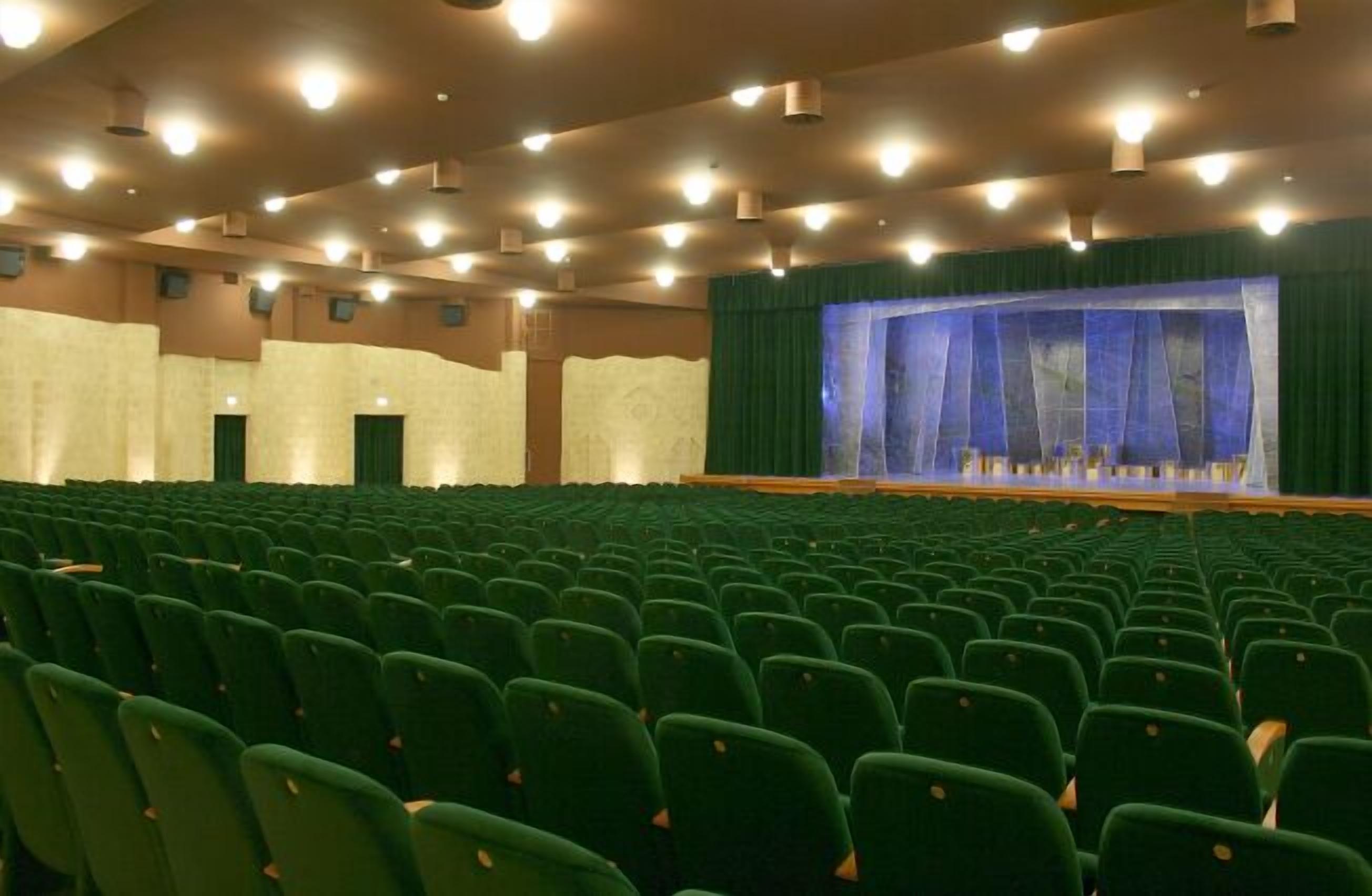 Un auditorio amplio con hileras de sillas verdes, un escenario con un telón azul y varias luces en el techo.