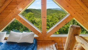 Uma cabana de madeira com uma cama e duas janelas, oferecendo vista para uma cadeia montanhosa coberta por floresta.