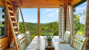 Uma cabana de madeira com mesa e cadeiras, uma escada e vista para uma cadeia montanhosa arborizada.