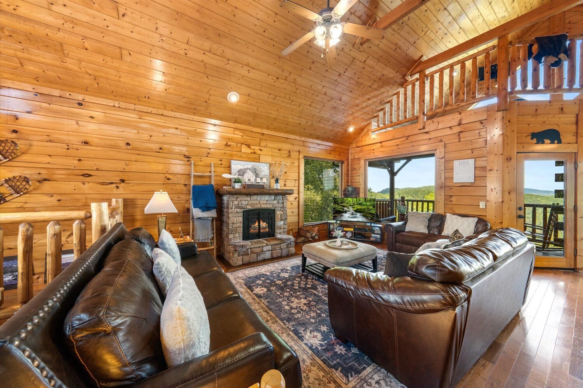 Un intérieur de chalet chaleureux avec une cheminée, des canapés en cuir, un plafond en bois et un balcon offrant une vue panoramique.