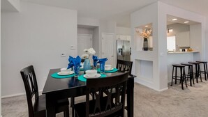 Un coin repas avec une table noire dressée pour quatre personnes, des murs blancs et une cuisine en arrière-plan.