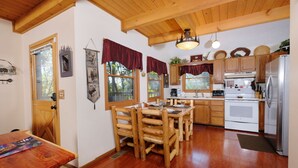 A wooden dining table with chairs, a kitchen with wooden cabinets and appliances, and a window with a view of trees. - Smoky Mountain High: Where the Mountains Lift You (Gatlinburg)