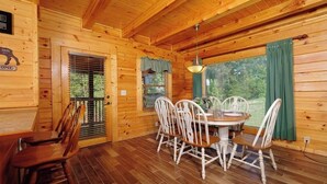 A wooden cabin with a dining area featuring a round table and white chairs, a wooden floor, and a view through the window.