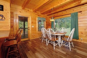 Una cabaña de madera con comedor que incluye una mesa redonda y sillas blancas, piso de madera y vista por la ventana.