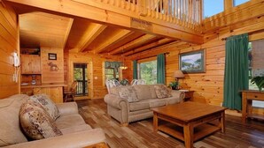A cozy cabin interior with a wooden floor, two sofas, a coffee table, a dining table, and a kitchen area.