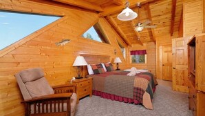 A cozy bedroom with a sloped ceiling, wooden walls, a bed with multiple pillows, a nightstand, a chair, and a window.