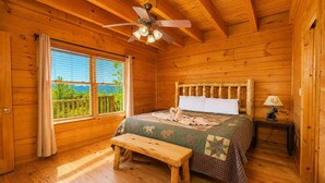 A wooden cabin bedroom with a bed, bench, ceiling fan, and a window with a view. - Deer Lodge: Putt, Play & Take in the Views (Pigeon Forge)