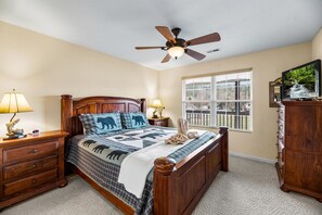 Quarto com cama de madeira, duas mesas de cabeceira com luminárias, ventilador de teto, janela grande com persianas e televisão sobre um armário de madeira.