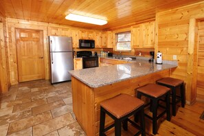 A wooden cabin kitchen with a granite countertop, a refrigerator, a microwave, a stove, and bar stools.