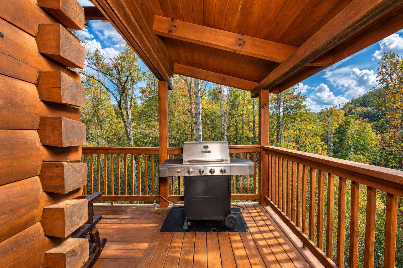 A covered outdoor deck with a barbecue grill, wooden railings, and a view of trees.