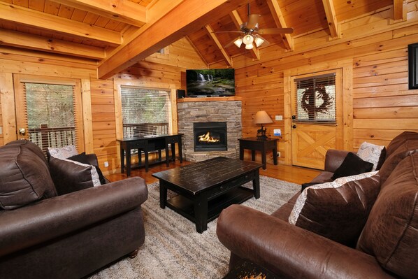 A log cabin interior with a fireplace, leather sofas, a coffee table, and a television mounted above the fireplace.