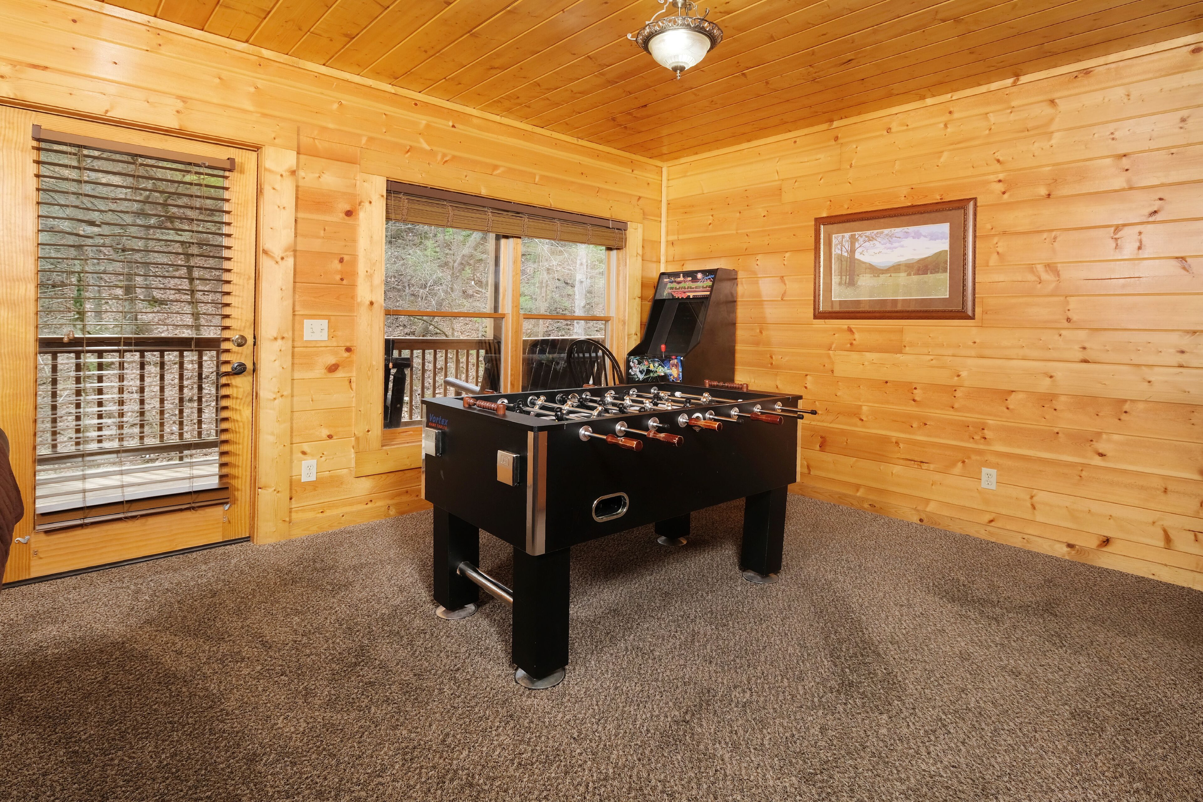 A wooden-floored room with a foosball table, a window with blinds, and a framed picture on the wall.