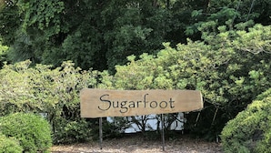Ein Holzschild mit dem Schriftzug 'Sugarfoot', platziert in einem Gartenbereich mit verschiedenen Sträuchern und Bäumen im Hintergrund.