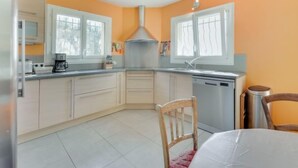 Una cocina compacta con armarios blancos, un lavamanos de acero inoxidable y un lavavajillas. Hay dos ventanas que proporcionan luz natural, y una silla de madera es visible en primer plano.