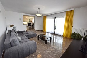 A modern living room with a grey sectional sofa, a coffee table, and a TV. There is a kitchen in the background with a yellow curtain.