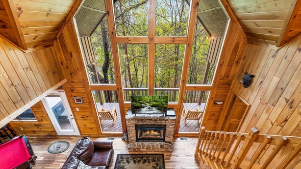 A cabin with a stone fireplace, wooden deck, and a large window overlooking a forest. - Raccoons' Rest: Kick Back in the Smokies (Pigeon Forge)